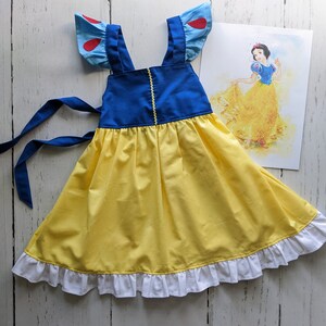 Puede incluir: Un vestido amarillo y azul con volantes blancos alrededor del dobladillo. El vestido tiene tirantes azules y detalles rojos en las mangas. El vestido es un disfraz de Blancanieves.
