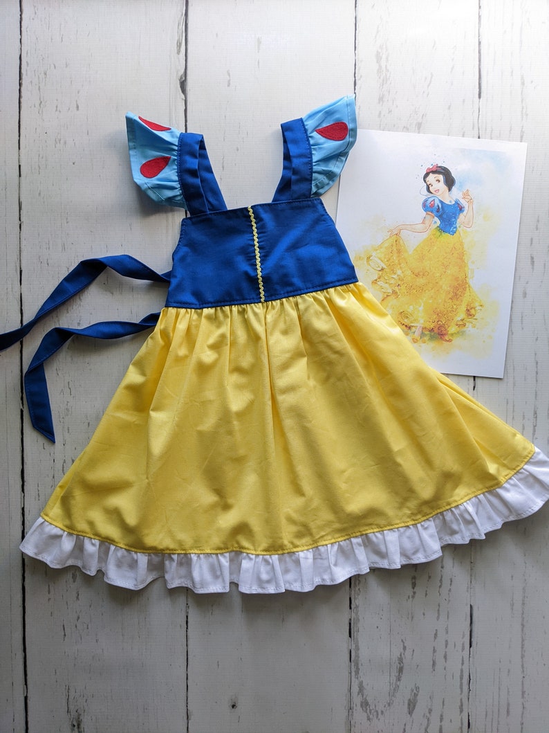 Puede incluir: Un vestido amarillo y azul con volantes blancos alrededor del dobladillo. El vestido tiene tirantes azules y detalles rojos en las mangas. El vestido es un disfraz de Blancanieves.