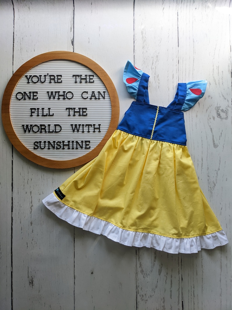 Puede incluir: Un vestido amarillo y azul con volantes blancos y detalles rojos. El vestido tiene un corpi&ntilde;o azul y una falda amarilla. El vestido tiene un letrero que dice "You're the one who can fill the world with sunshine".