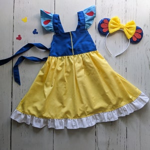 Puede incluir: Un vestido amarillo con tirantes azules y ribete de volantes blancos. El vestido tiene mangas abullonadas azules y rojas. Una diadema con un lazo rojo y amarillo con detalles de purpurina roja.