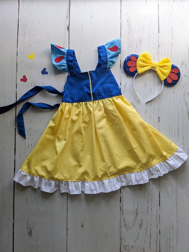 Puede incluir: Un vestido amarillo con tirantes azules y ribete de volantes blancos. El vestido tiene mangas abullonadas azules y rojas. Una diadema con un lazo rojo y amarillo con detalles de purpurina roja.