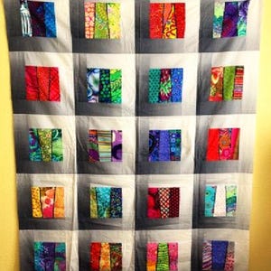May include: A quilt with a patchwork design featuring colorful fabric strips in various patterns. The quilt is made up of 16 squares, each with a different pattern. The quilt is hanging on a wall.