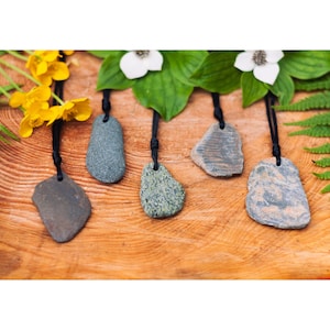May include: Five natural stone pendants suspended on thin black cords. Each pendant features a unique, irregularly shaped rock in varying shades of gray, green, and brown, with visible textures and patterns, each drilled with a hole for the cord.