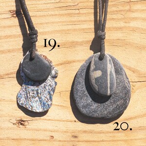 May include: Two stone pendants on black cords. One pendant features a dark gray stone atop a piece of iridescent shell. The other pendant has a gray stone with a white stripe on top of a larger gray stone. The background is a wooden surface.
