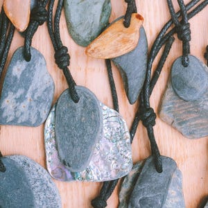 May include: A collection of stone pendant necklaces with black cords. The pendants vary in shape and color, including gray, brown, and iridescent shell pieces. The necklaces are displayed on a light wooden surface.