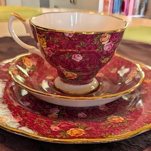 May include: A vintage tea set featuring a burgundy cup, saucer, and plate, adorned with a floral pattern in shades of pink, yellow, and white. The set has gold trim, adding a touch of elegance to the design. A classic tea party set.