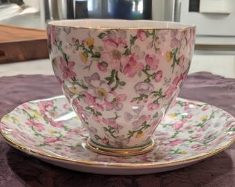 May include: A porcelain teacup and saucer set featuring a white base with a dense floral pattern of pink, yellow, and light purple blossoms on green stems. Both the cup and the saucer have scalloped edges trimmed with a thin gold metallic band.