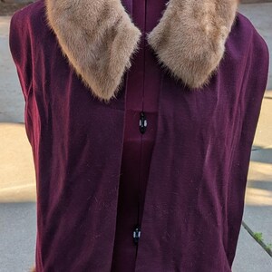 May include: A vintage burgundy cape with a fur collar and trim. The cape is displayed on a dress form, showcasing its open front design. The fur appears to be a light brown color, contrasting with the deep purple of the cape.