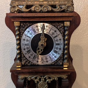 May include: Antique wall clock with a dark wood frame and ornate brass detailing. The clock face is silver with black Roman numerals. Features a decorative top with figures and a pendulum with brass weights.