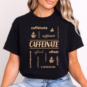 Może przedstawiać: Czarny t-shirt ze słowem "CAFFEINATE" w złotym kolorze. Projekt zawiera ilustracje filiżanek i ziaren kawy, a słowo "caffeinate" jest powtórzone mniejszą czcionką. To codzienna koszulka.