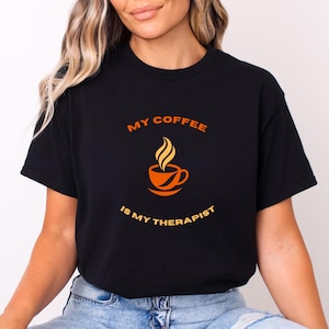 Può includere: T-shirt nero con la scritta "MY COFFEE IS MY THERAPIST" in arancione. La maglietta presenta una grafica di una tazza di caffè arancione con vapore che sale. È un capo di abbigliamento casual.