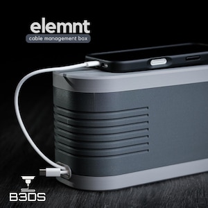 May include: A gray and white cable management box with a black smartphone on top. The box has a rectangular shape with a ridged design on the sides. A white charging cable is plugged into the phone and the box. The text "elemnt cable management box" is visible.