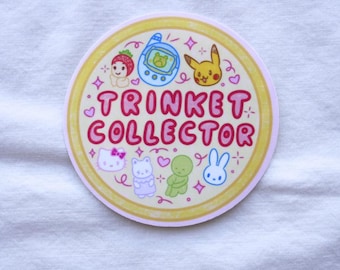 May include: A round sticker with a pink and yellow border featuring cartoon characters and the text "TRINKET COLLECTOR" in pink. The sticker includes images of a strawberry, a handheld game, Pikachu, Hello Kitty, a bear, a green alien, and a bunny.