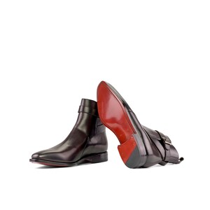 K&ouml;nnte beinhalten: Ein Paar dunkelbraune Leder-Ankle Boots mit spitzer Zehenpartie und Riemen mit Schnalle. Die Sohlen sind rot. Die Stiefel werden auf wei&szlig;em Hintergrund pr&auml;sentiert.