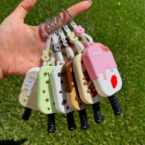 May include: A collection of six popsicle-shaped keychains attached to a silver metal ring. Each keychain features a unique color, including pink, cream, brown, and light green, with small decorative details and the word Play printed on the pink one.