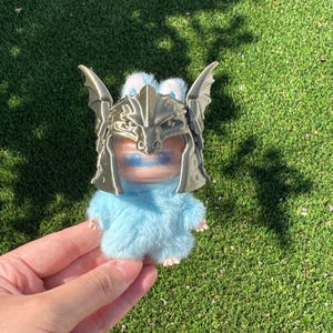 May include: A small, fuzzy light blue creature figurine wearing a metallic gold-colored dragon-shaped helmet. The creature has a smooth face with blue eyes and a soft, textured body. The helmet features detailed dragon wings and scales.