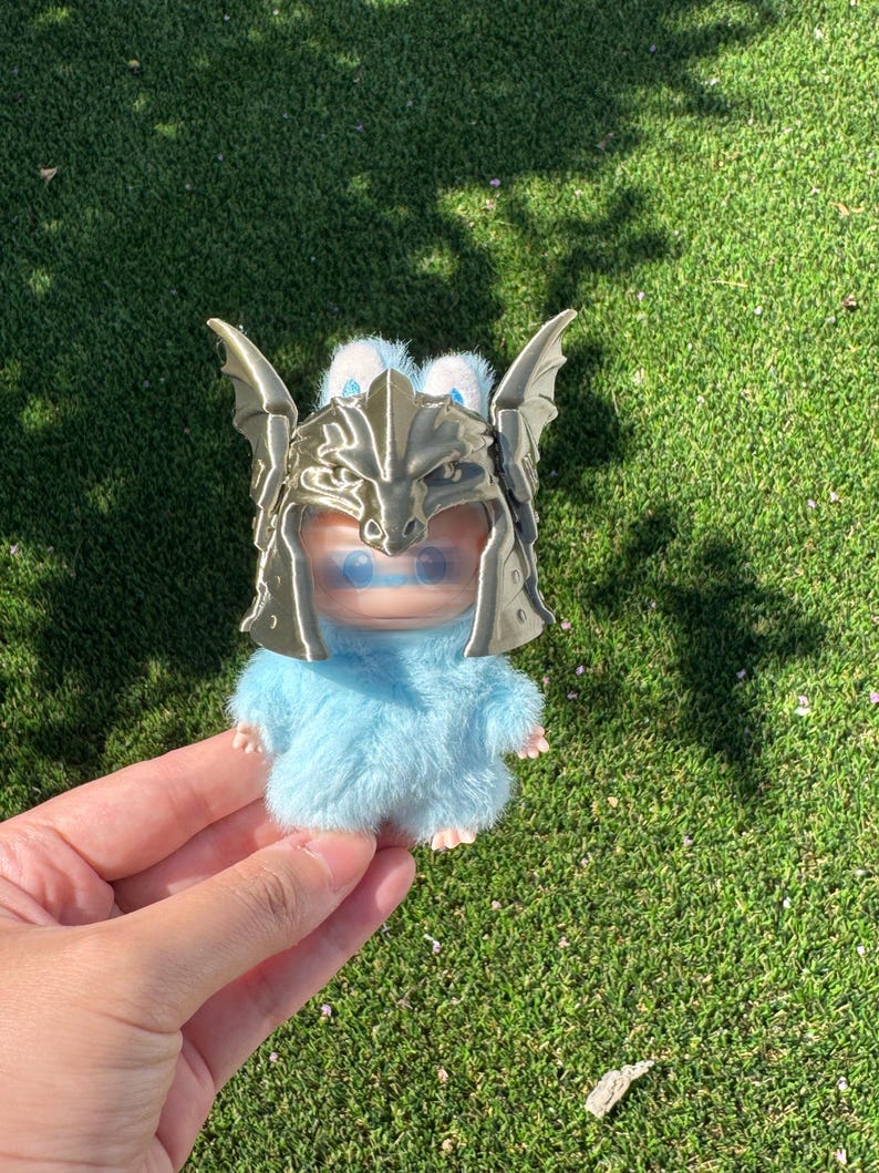May include: A small, fuzzy light blue creature figurine wearing a metallic gold-colored dragon-shaped helmet. The creature has a smooth face with blue eyes and a soft, textured body. The helmet features detailed dragon wings and scales.