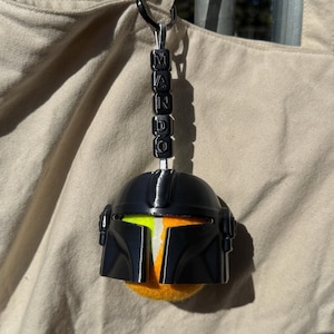 Puede incluir: Un llavero de casco mandaloriano negro con detalles en naranja y amarillo. El llavero tiene la palabra "MANDO" escrita con cuentas negras. El casco est&aacute; unido a un anillo de metal negro.