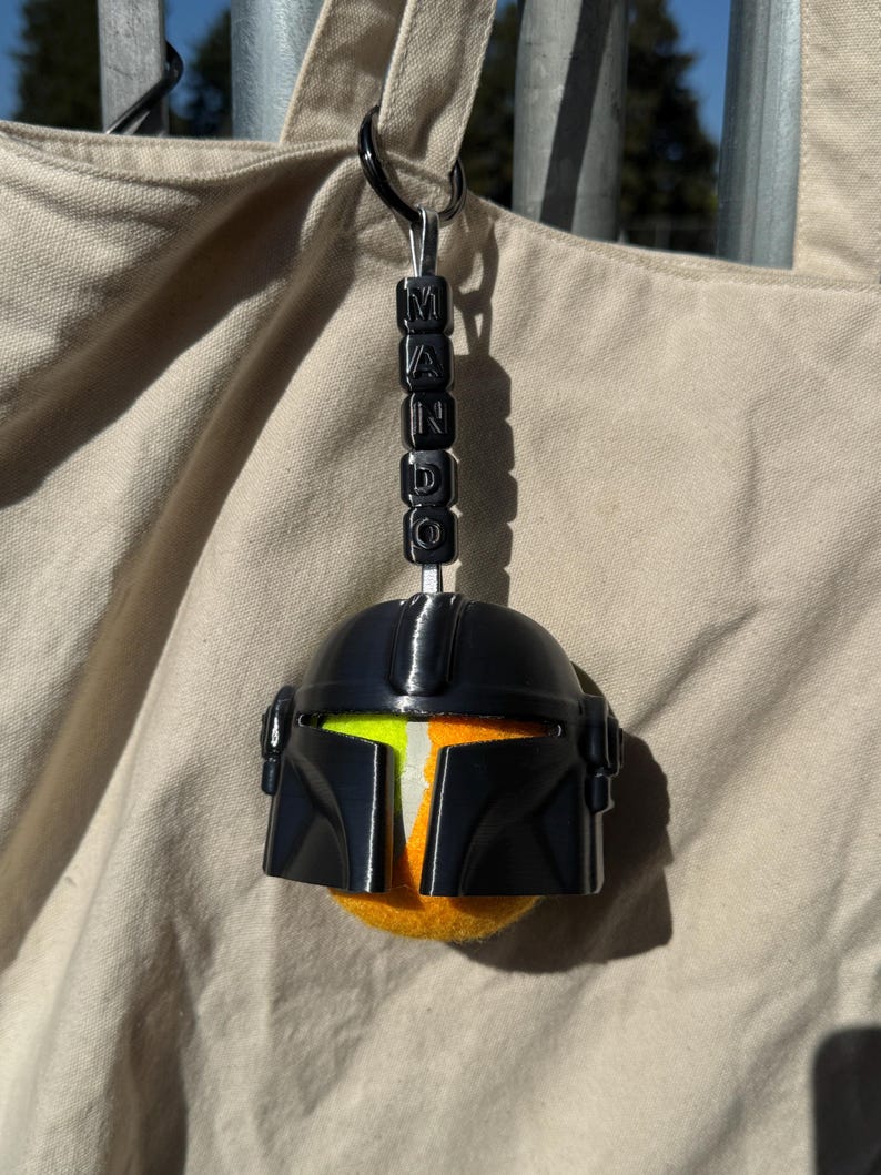Puede incluir: Un llavero de casco mandaloriano negro con detalles en naranja y amarillo. El llavero tiene la palabra "MANDO" escrita con cuentas negras. El casco est&aacute; unido a un anillo de metal negro.