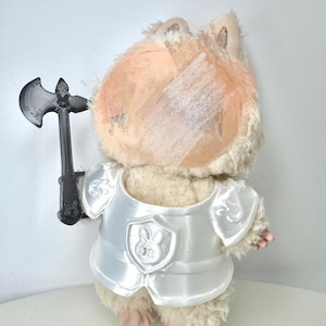 Może przedstawiać: Pluszowa zabawka chomika ubranego w błyszczącą białą zbroję rycerską z tarczą z emblematem królika. Chomik trzyma czarną plastikową siekierę. Zabawka ma jasnobrązowe futro i bandaż na głowie.