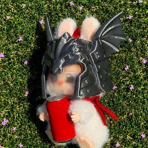 May include: A white plush rabbit figurine wearing a dark gray, metallic-finish dragon-shaped helmet with wings. The rabbit has a red scarf around its neck and is holding a small, solid red cylindrical can in its front paws.
