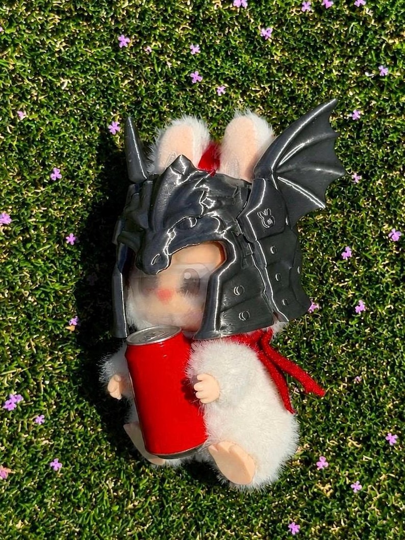 May include: A white plush rabbit figurine wearing a dark gray, metallic-finish dragon-shaped helmet with wings. The rabbit has a red scarf around its neck and is holding a small, solid red cylindrical can in its front paws.