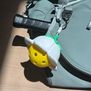 May include: A pickleball keychain featuring a yellow perforated ball encased in a white flower-shaped holder with green stem details, attached to a silver metal ring and a white plastic clip, hanging from a sage green sports bag.