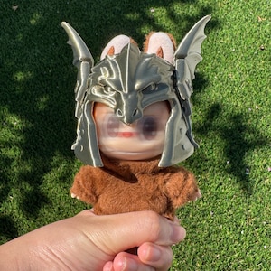 May include: A small plush figure with brown fur wearing a metallic gold-colored dragon-shaped helmet. The helmet features prominent wing-like side panels and a detailed dragon head design covering the forehead area of the plush character.