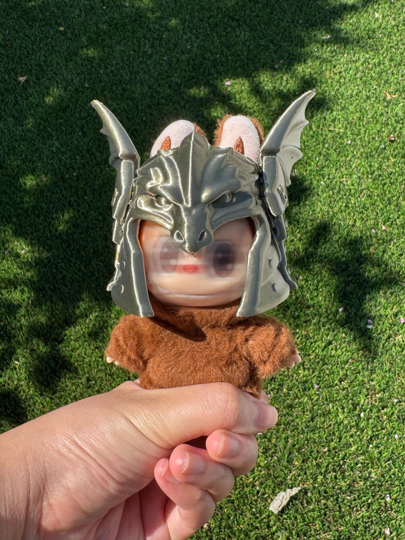 May include: A small plush figure with brown fur wearing a metallic gold-colored dragon-shaped helmet. The helmet features prominent wing-like side panels and a detailed dragon head design covering the forehead area of the plush character.