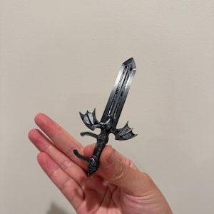 May include: A small, dark gray, 3D-printed sword featuring a detailed hilt shaped like a bat with outstretched wings. The blade is straight with a central ridge, and the handle has a textured grip, ending in a rounded pommel at the base.