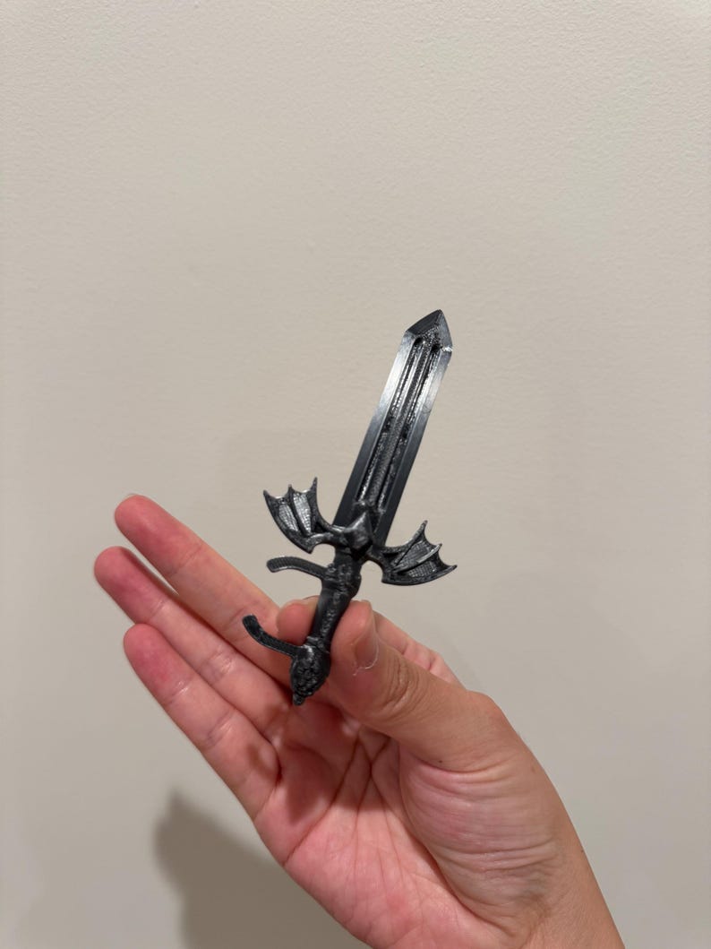 May include: A small, dark gray, 3D-printed sword featuring a detailed hilt shaped like a bat with outstretched wings. The blade is straight with a central ridge, and the handle has a textured grip, ending in a rounded pommel at the base.