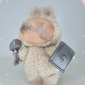 Op de afbeelding: Een pluizig, crèmekleurig pluche speelgoedfiguur, met een zilveren hamer en schild. Het speelgoed heeft een lichtbruin gezicht en staat tegen een zachte, vage achtergrond met een gloeiend effect. Het speelgoed is een grillige weergave van een krijger.
