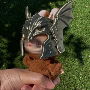May include: A small brown fuzzy plush figure wearing a metallic bronze-colored dragon-themed helmet. The helmet features sculpted dragon scales, a pointed snout design, and large, textured dragon wings extending from the sides of the headpiece.