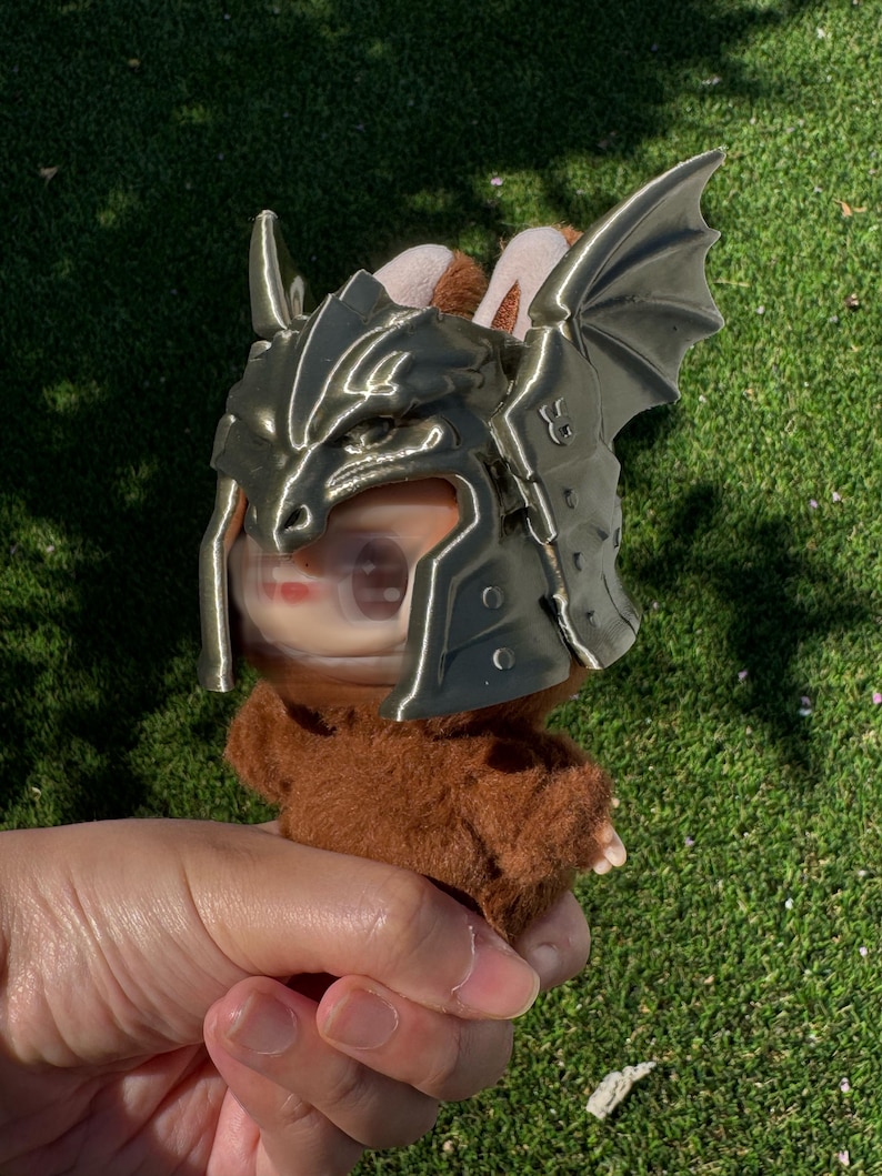 May include: A small brown fuzzy plush figure wearing a metallic bronze-colored dragon-themed helmet. The helmet features sculpted dragon scales, a pointed snout design, and large, textured dragon wings extending from the sides of the headpiece.