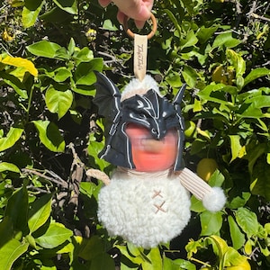 May include: A plush white round ornament featuring a black dragon-style helmet with wings, a beige striped scarf, and a small beige tag with the text Twinkle Twinkle. The ornament hangs from a brown wooden ring, showcasing a soft, fuzzy texture.