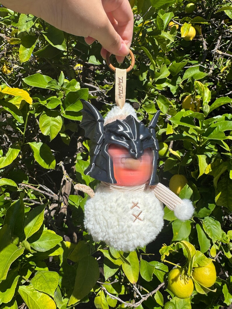 May include: A plush white round ornament featuring a black dragon-style helmet with wings, a beige striped scarf, and a small beige tag with the text Twinkle Twinkle. The ornament hangs from a brown wooden ring, showcasing a soft, fuzzy texture.