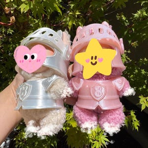 Op de afbeelding: Twee pluche speelgoed gekleed als ridders. E&eacute;n is cr&egrave;mekleurig met een zilveren helm en harnas, de andere is roze met een bijpassende roze helm en harnas. Beide hebben een schildontwerp op hun borst. De achtergrond is groen gebladerte.