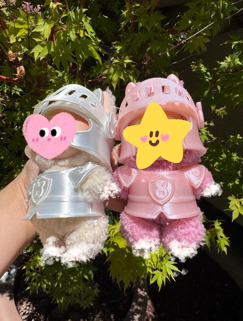 Op de afbeelding: Twee pluche speelgoed gekleed als ridders. E&eacute;n is cr&egrave;mekleurig met een zilveren helm en harnas, de andere is roze met een bijpassende roze helm en harnas. Beide hebben een schildontwerp op hun borst. De achtergrond is groen gebladerte.