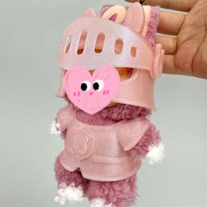 Op de afbeelding: Een roze pluche speelgoed in ridderharnas, met helm en borstpantser. Het speelgoed heeft een pluizig, roze lichaam en witte accenten op de poten. Een goudkleurige sleutelhanger is aan de bovenkant bevestigd. Ongeveer 15 cm hoog.