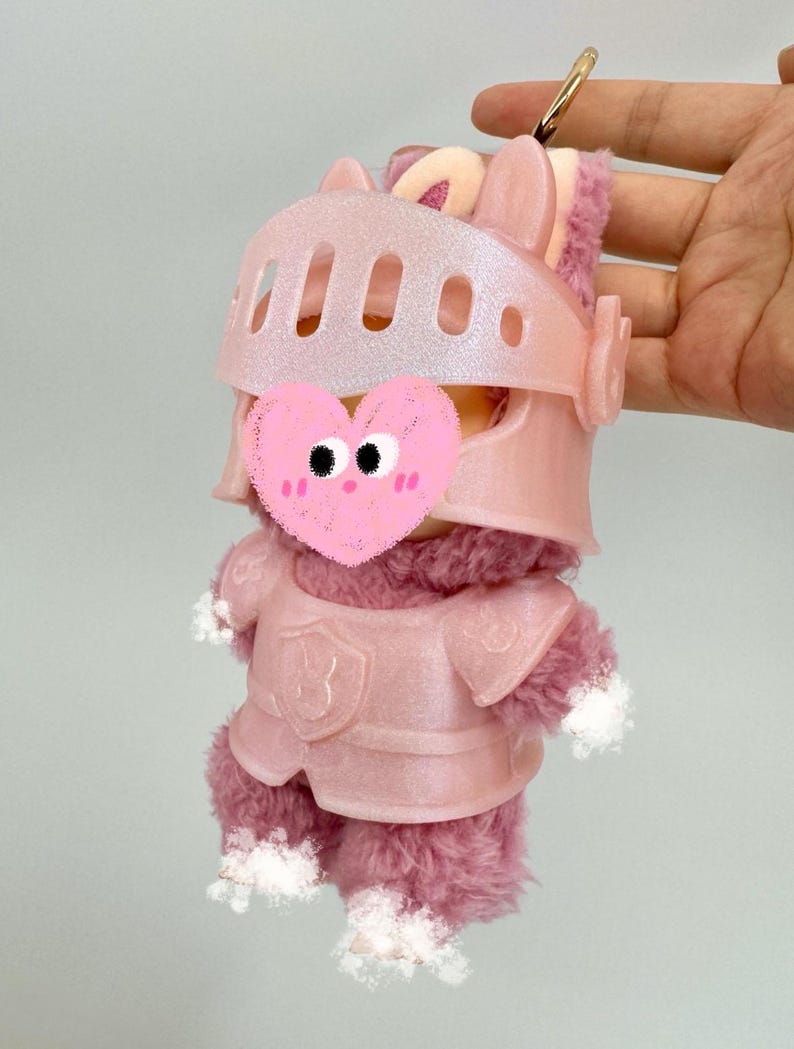 Op de afbeelding: Een roze pluche speelgoed in ridderharnas, met helm en borstpantser. Het speelgoed heeft een pluizig, roze lichaam en witte accenten op de poten. Een goudkleurige sleutelhanger is aan de bovenkant bevestigd. Ongeveer 15 cm hoog.