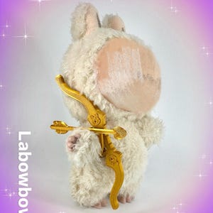 Op de afbeelding: Een pluche, gebroken witte lammetjesknuffel met een gouden pijl en boog. Het lam heeft een getextureerde vacht en roze accenten op de oren en het gezicht. Pijl en boog zijn ingewikkeld ontworpen. De tekst "Labowbow" staat verticaal aan de linkerkant.