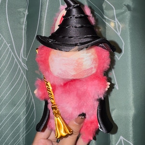 Może przedstawiać: Różowa pluszowa zabawka przebrana za czarownicę, z czarnym kapeluszem i peleryną. Zabawka trzyma małą złotą miotłę. Plusz ma puszystą fakturę i fantazyjny design, idealny na Halloween lub dekoracje w stylu fantasy.