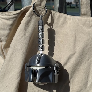Puede incluir: Un llavero de casco mandaloriano plateado con la palabra "MANDO" deletreada en cuentas de letras individuales. El llavero est&aacute; sujeto a un anillo plateado y cuelga frente a una bolsa de mano beige.