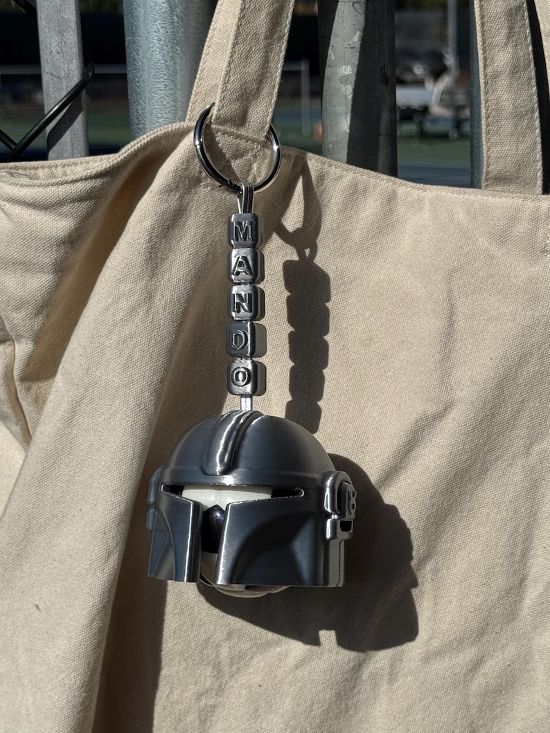 Puede incluir: Un llavero de casco mandaloriano plateado con la palabra "MANDO" deletreada en cuentas de letras individuales. El llavero est&aacute; sujeto a un anillo plateado y cuelga frente a una bolsa de mano beige.
