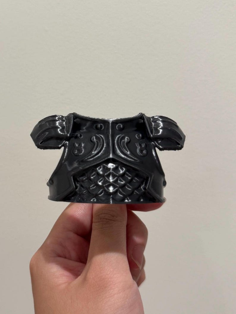 May include: A black, molded plastic chest piece armor accessory featuring a glossy finish, shoulder pauldrons, and a textured scale-like pattern on the lower front section, designed with a rigid structure and symmetrical decorative carvings.