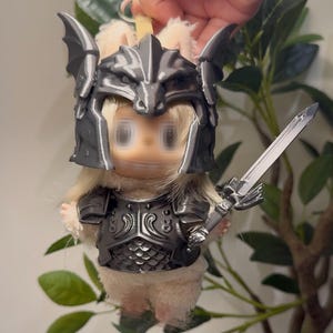 May include: A small plush figure with light beige fur wearing a metallic dark gray 3D-printed dragon-themed helmet and matching chest armor. The figure holds a small metallic gray sword in its hand and features a keychain attachment at the top.