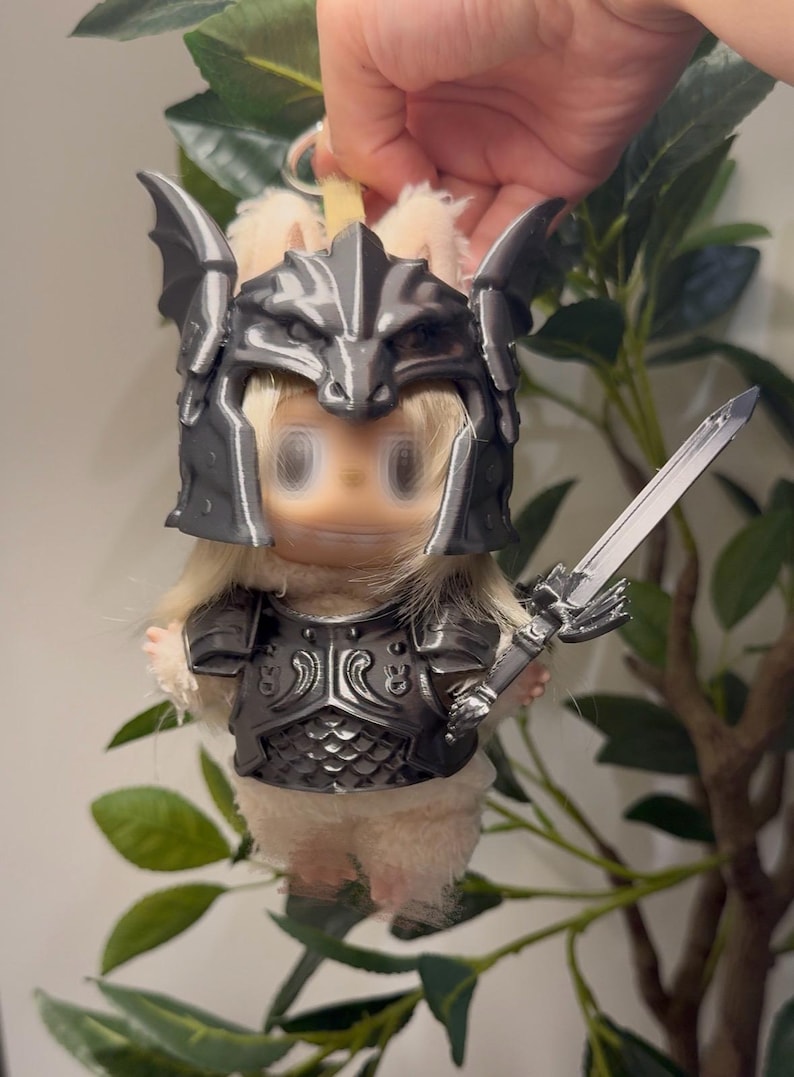 May include: A small plush figure with light beige fur wearing a metallic dark gray 3D-printed dragon-themed helmet and matching chest armor. The figure holds a small metallic gray sword in its hand and features a keychain attachment at the top.
