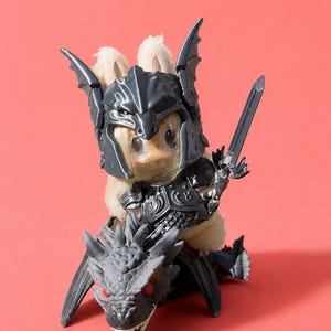 May include: A fuzzy, cream-colored rabbit figurine wearing a dark gray, detailed knight's helmet and chest armor. The rabbit holds a small gray sword in one hand and is mounted on a dark gray, scaly dragon figure with red eyes and sharp teeth.