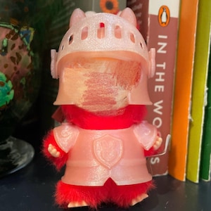 Puede incluir: Un pequeño juguete de peluche de un caballero con armadura y casco rosas, con un cuerpo y brazos rojos y peludos. El caballero tiene un escudo en el pecho y una espada pequeña.