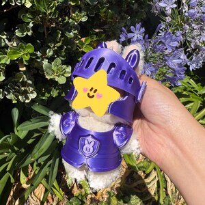 Op de afbeelding: Een wit pluche konijn met een paarse ridderhelm en -pantser. Het pantser heeft een konijnenwapen. Het konijn wordt vastgehouden voor een achtergrond van groen gebladerte en paarse bloemen.